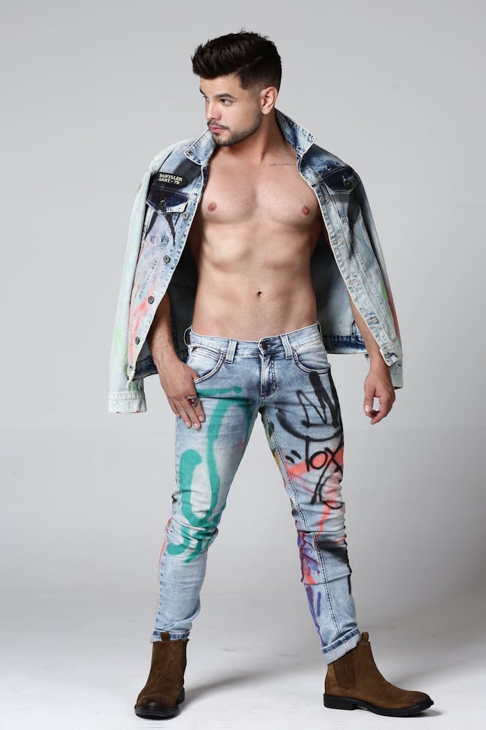 Shirtless male model showcasing fashion-forward denim with graffiti art. Studio shot on white background.
